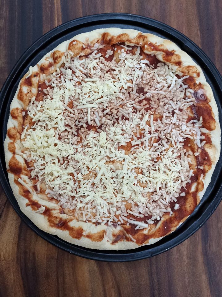 Pizza cruda con queso rallado sobre la salsa, en su bandeja negra sobre una mesa de madera, antes de hornear